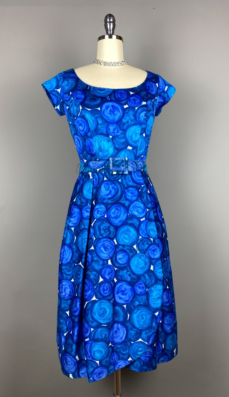 May include: A blue and white vintage dress with a circle pattern. The dress has a scoop neckline, short sleeves, and a fitted bodice. It is belted at the waist and has a full skirt.