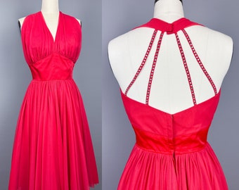 May include: A bright pink vintage dress with a halter neckline and a full skirt. The dress has a unique back design with a criss-cross strap detail.