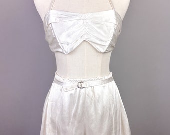 May include: White satin two-piece set with a halter top and shorts. The top has a sweetheart neckline and the shorts have a belt with a silver buckle. The set is accessorized with a red beaded necklace.