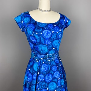 May include: A blue and white vintage dress with a circle pattern. The dress has a scoop neck, short sleeves, and a fitted bodice. It has a belt at the waist and a full skirt.