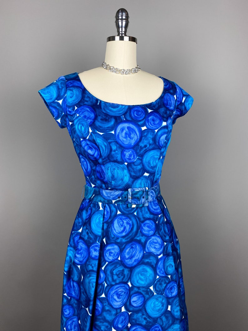 May include: A blue and white vintage dress with a circle pattern. The dress has a scoop neck, short sleeves, and a fitted bodice. It has a belt at the waist and a full skirt.
