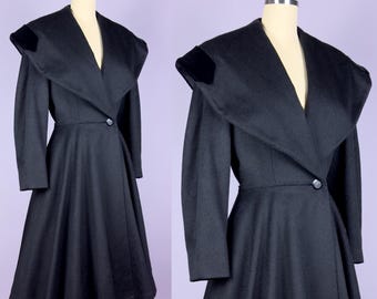Puede incluir: Un abrigo negro vintage con un cuello ancho y una falda acampanada. El abrigo presenta un cierre de un solo botón en la cintura y detalles de terciopelo en los hombros. El abrigo se exhibe en un maniquí.