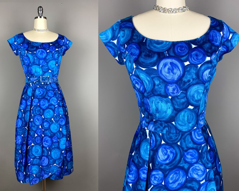May include: A blue and white vintage dress with a circle pattern. The dress has a scoop neck, short sleeves, and a fitted bodice. It has a full skirt and a belt at the waist.