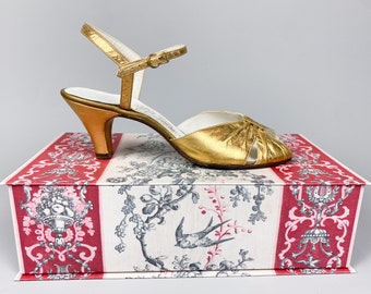 Puede incluir: Una sandalia de tacón de cuero dorado con una correa de tobillo. La sandalia está sentada en una caja con un patrón floral.