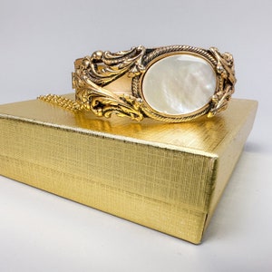 May include: A gold-tone bracelet with an ornate design and a large, white mother-of-pearl oval centerpiece.