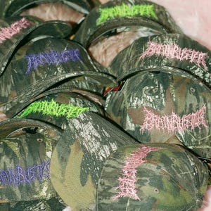 Puede incluir: Un montón de gorras de béisbol de camuflaje con la palabra "Brainbuster" bordada en diferentes colores, incluyendo rosa, verde y morado.
