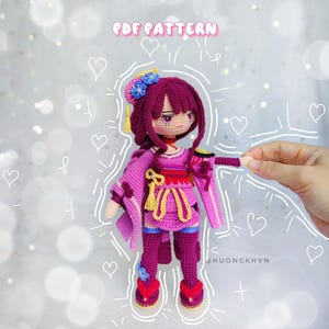 Puede incluir: Patrón de muñeca amigurumi de crochet en formato PDF. La muñeca lleva un kimono rosa y amarillo con una flor morada en el pelo. La muñeca está de pie sobre un fondo blanco con corazones y líneas dibujadas a su alrededor.