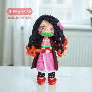 May include: A handmade crochet doll with black hair, pink eyes, and a green mouth guard. The doll wears a pink dress, brown coat, and red shoes. The doll's hair has orange accents. The text "DOWNLOAD ENGLISH PATTERN" is in the upper left corner.
