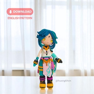 May include: A handmade crochet doll with a teal and blue color scheme. The doll has a detailed outfit with a white top, teal skirt, and black boots with yellow soles. The doll has blue hair and yellow eyes. The image includes the text "DOWNLOAD ENGLISH PATTERN".