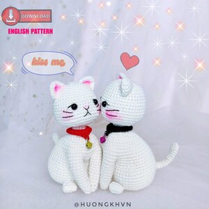 May include: Two white crocheted cat plushies with pink cheeks and black eyes. The cat on the left is wearing a red collar with a gold bell. The cat on the right is wearing a black collar with a purple bell. The cats are kissing and there is a speech bubble above them that says "kiss me".
