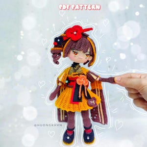 Könnte beinhalten: Gehäkelte Amigurumi-Puppe mit braunen Haaren, einer roten Blume auf dem Kopf und einem gelben und braunen Kleid. Die Puppe trägt schwarze Schuhe mit weißen Akzenten. Die Puppe ist von Herzen umgeben und der Text "PDF PATTERN" befindet sich oben im Bild.