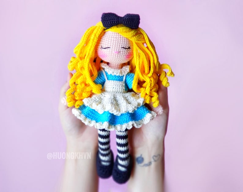 May include: A crocheted Alice in Wonderland doll with blonde hair, a blue and white dress, and black and white striped stockings. The doll is wearing a black bow and has a closed-eye smile.