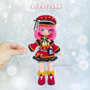 May include: A crocheted doll with pink hair, a red and black dress, and a black and yellow hat. The doll is wearing a monocle and has a red and yellow bow on its chest. The doll is standing on a white background with a white outline around it. The text "PDF PATTERN" is at the top of the image. The text "@HUONGKHVN" is at the bottom of the image.