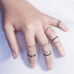 Könnte beinhalten: Eine Hand trägt einen Satz dünner, zarter Ringe in Gold, Silber und Schwarz. Die Ringe sind auf mehreren Fingern übereinander gelegt.