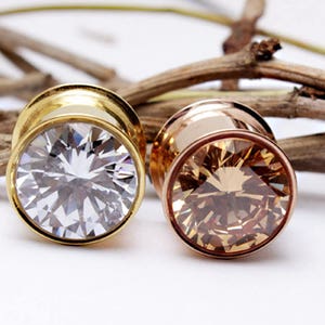 May include: Two pairs of ear gauges with jeweled accents. The gauge on the left is gold with a clear stone, and the gauge on the right is rose gold with a brown stone.