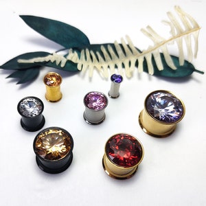 Könnte beinhalten: Eine Sammlung farbenfroher Ohr-Gauges mit großen, facettierten Edelsteinen. Die Gauges sind in verschiedenen Farben erhältlich, darunter klar, bernsteinfarben, lila und rot, gefasst in Gold, Silber und schwarzem Metall. Dekorative Elemente im Hintergrund.