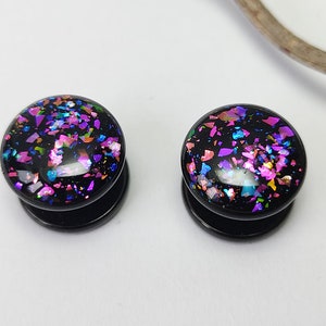 Può includere: Coppia di dilatatori per orecchie in acrilico nero con glitter iridescente incorporato nella superficie. Il glitter è un mix di rosa, blu e oro.