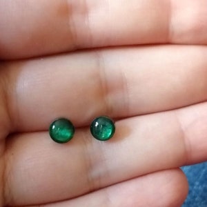 Könnte beinhalten: Ein Paar kleine, runde, grüne Ohrringe. Die Ohrringe sind aus Glas und haben eine glänzende, metallische Oberfläche.