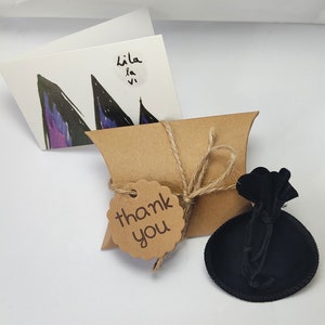 May include: A small brown paper gift bag with a thank you tag tied with twine. The bag is sitting on a white surface next to a black velvet pouch.