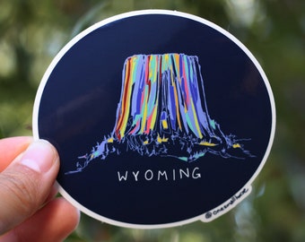 May include: A round sticker with a dark blue background featuring a colourful illustration of Devils Tower National Monument in Wyoming. The text "WYOMING" is printed below the illustration.