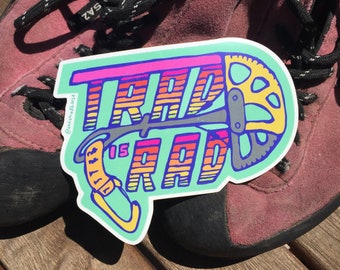May include: A turquoise and pink sticker with the text "TRAD IS RAD" in rainbow colours. The sticker features a stylised illustration of a climbing hold and a gear.