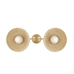 May include: A double-headed wall sconce with woven wicker shades and gold metal accents. The shades are round and have a slight curve to them. The sconce has two light bulbs.