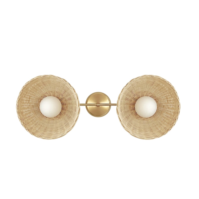 May include: A double-headed wall sconce with woven wicker shades and gold metal accents. The shades are round and have a slight curve to them. The sconce has two light bulbs.