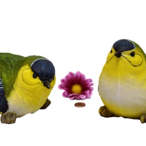Könnte beinhalten: Zwei Keramik-Vogelfiguren, eine grün und blau mit gelbem Bauch, die andere gelb mit weißem Bauch und blauen Flügeln. Beide Vögel sitzen auf einer weißen Oberfläche.