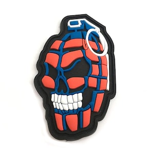 Puede incluir: Un parche de granada en forma de calavera rojo y azul con una mecha blanca. El parche está hecho de goma y tiene un borde negro.
