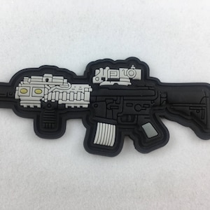 Könnte beinhalten: Schwarz-weiß-Patch einer Sturmgewehrs mit Zielfernrohr. Der Patch ist aus einem gummiartigen Material.