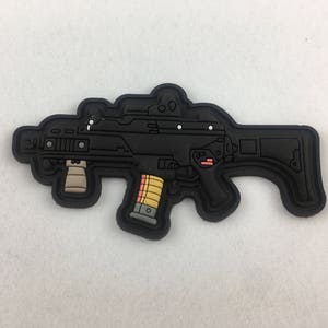May include: Black and white patch of a submachine gun with a magazine inserted. The patch is made of fabric and has a textured surface.