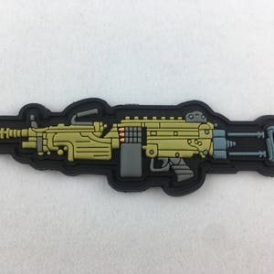May include: A black and gold machine gun patch with a detailed design. The patch is made of rubber and has a black border.