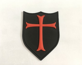 Puede incluir: Parche negro en forma de escudo con una cruz roja en el centro. El parche está hecho de un material texturizado.