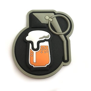 Puede incluir: Un parche verde y negro con forma de granada con un vaso de cerveza en su interior. El vaso de cerveza es naranja con espuma blanca en la parte superior.