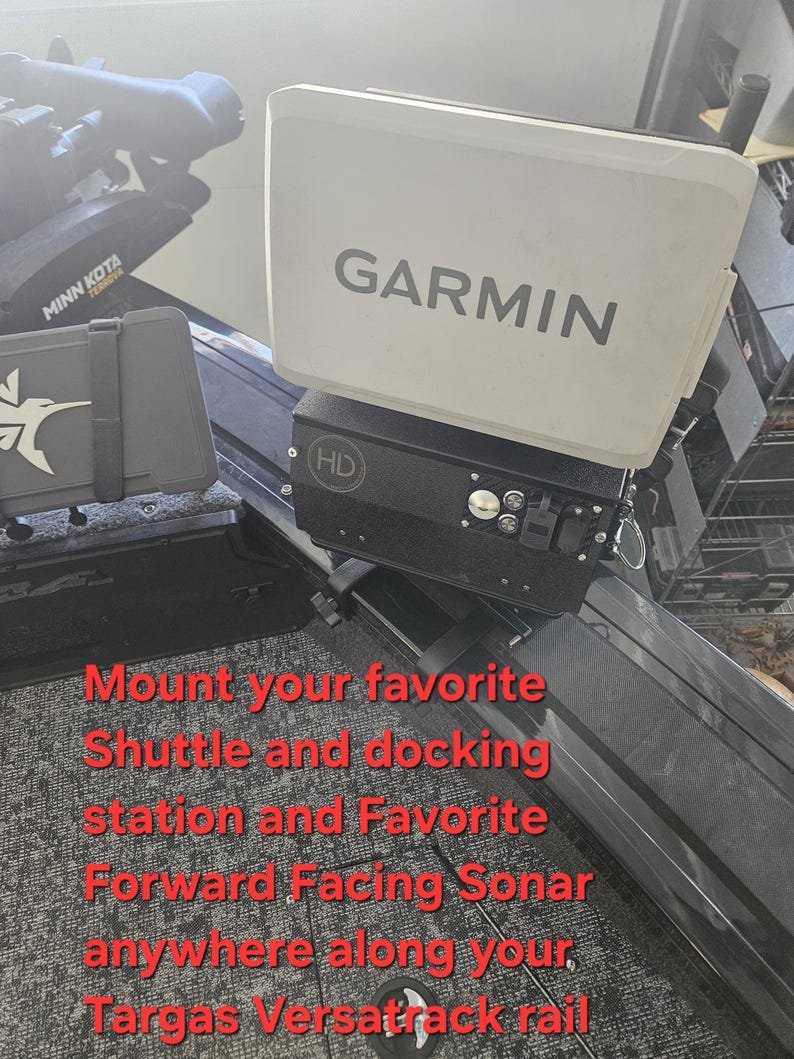 Puede incluir: Un buscador de peces Garmin blanco con el texto "GARMIN" en gris. El buscador de peces est&aacute; montado sobre una base negra con botones y una peque&ntilde;a pantalla. La imagen tambi&eacute;n incluye texto que dice "Monte su estaci&oacute;n de acoplamiento y sonar de avance favorito en cualquier lugar a lo largo de su riel Targas Versatrack."
