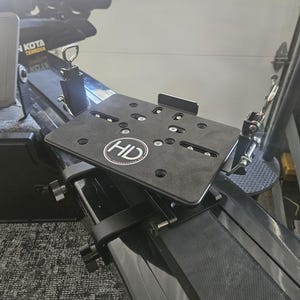 Puede incluir: Una placa de montaje de metal negro con las letras "HD" en un c&iacute;rculo blanco est&aacute; unida a un componente de barco negro. La placa tiene m&uacute;ltiples agujeros y herrajes de montaje. La imagen muestra un primer plano del sistema de montaje.