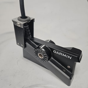 Puede incluir: Transductor de sonda Garmin negro con un cable negro. El transductor tiene un soporte metálico y una sección acanalada. La palabra "GARMIN" está impresa en blanco en el lateral del dispositivo.