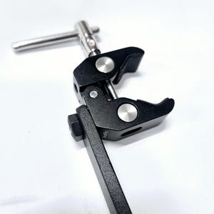 Puede incluir: Una abrazadera de metal negro con un mango plateado y una perilla plateada. La abrazadera est&aacute; unida a una varilla de metal negro.