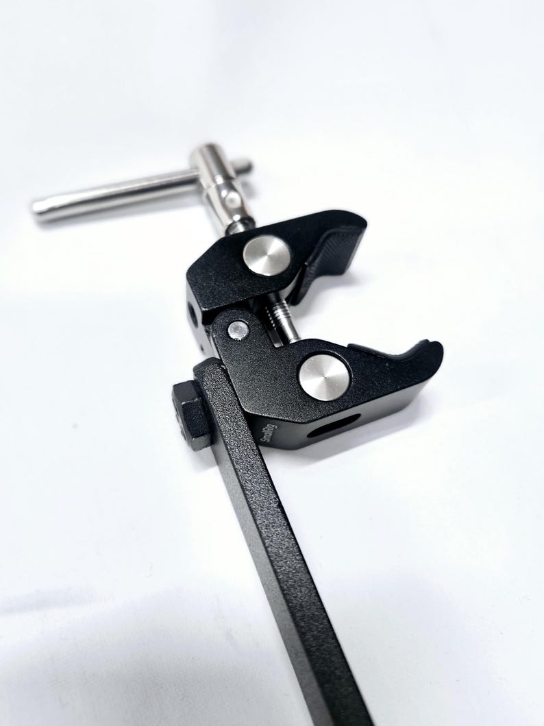 Puede incluir: Una abrazadera de metal negro con un mango plateado y una perilla plateada. La abrazadera est&aacute; unida a una varilla de metal negro.