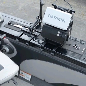 Puede incluir: Un buscador de peces Garmin y equipos relacionados est&aacute;n montados en un barco. El buscador de peces es blanco con el logotipo de Garmin. El barco tiene detalles en negro y gris, con un asiento blanco visible.