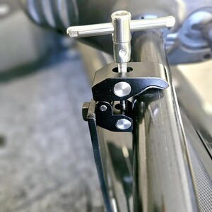 Puede incluir: Una abrazadera de metal negro con una perilla plateada y un mango en forma de T. La abrazadera est&aacute; unida a un tubo de metal negro.