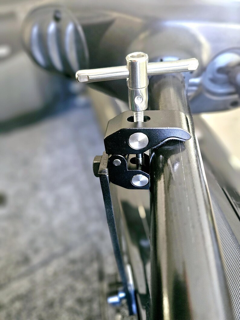 Puede incluir: Una abrazadera de metal negro con una perilla plateada y un mango en forma de T. La abrazadera est&aacute; unida a un tubo de metal negro.