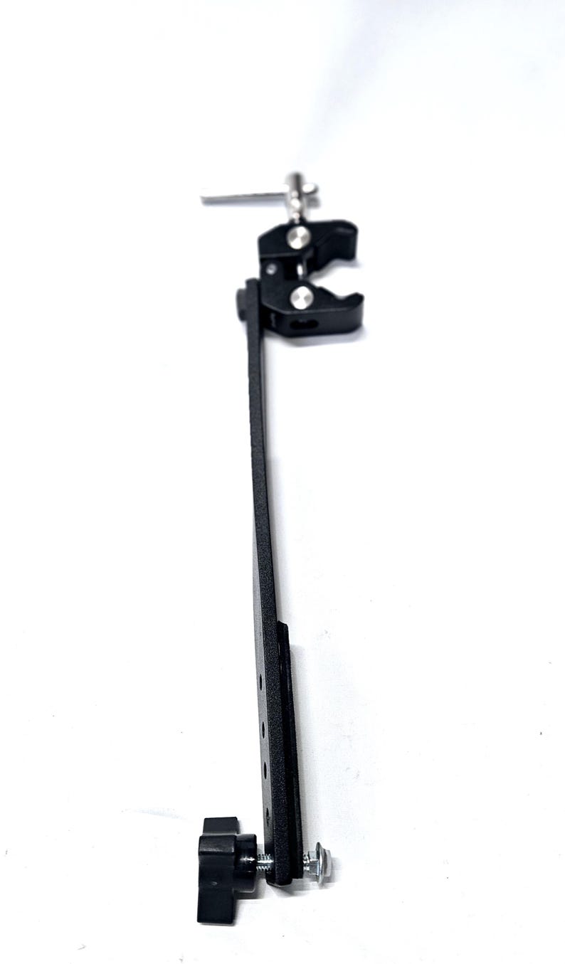 Puede incluir: Una abrazadera de metal negro con un brazo largo y una perilla en la parte inferior. La abrazadera tiene un tornillo plateado y una perilla plateada en la parte superior.