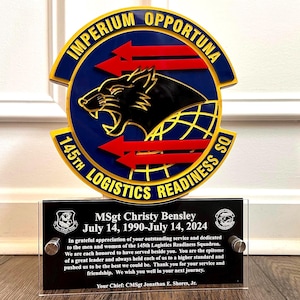 May include: A black and gold plaque with a blue and red emblem featuring a wolf and the text "Imperium Opportunia 145th Logistics Readiness SQ". The plaque has a black background with white text that reads "MSgt Christy Bensley July 14, 1990-July 14, 2024 In grateful appreciation of your outstanding service and dedicated to the men and women of the 145th Logistics Readiness Squadron. We are each honored to have served beside you. You are the epitome of a great leader and always held each of us to a higher standard and pushed us to be the best we could be. Thank you for your service and friendship. We wish you well in your next journey. Your Chief: CMSgt Jonathan E. Shores, Jr."