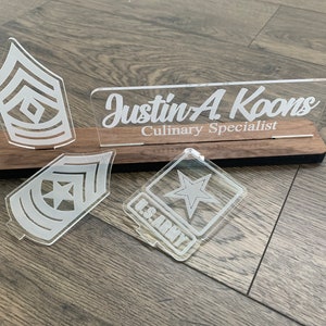 May include: A wooden plaque with a clear acrylic nameplate that reads "Justin A. Koons Culinary Specialist". There are three clear acrylic military insignia on the plaque, including a star and two rank insignia.