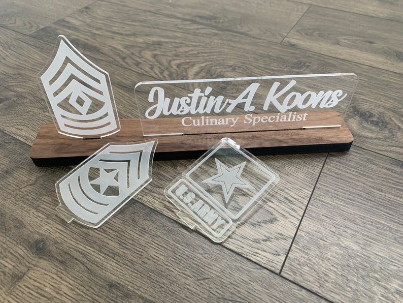 May include: A wooden plaque with a clear acrylic nameplate that reads "Justin A. Koons Culinary Specialist". There are three clear acrylic military insignia on the plaque, including a star and two rank insignia.