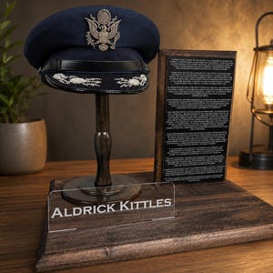 May include: A dark blue military-style service cap with a silver eagle emblem and black visor, displayed on a wooden stand. A clear acrylic plaque reads ALDRICK KITTLES, positioned in front of a wooden board with engraved text detailing officer duties.