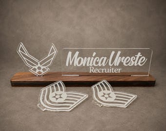 May include: A dark wood desk stand holding a clear acrylic nameplate with the text Monica Ureste Recruiter, an acrylic Air Force logo, and two separate acrylic Air Force rank insignia pieces placed in front of the wooden base.