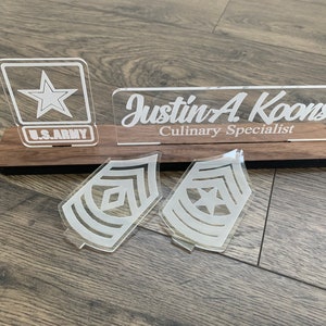 May include: A wooden plaque with a nameplate that reads "Justin A. Koons Culinary Specialist" and a U.S. Army logo. There are two white acrylic military rank insignia on the plaque.