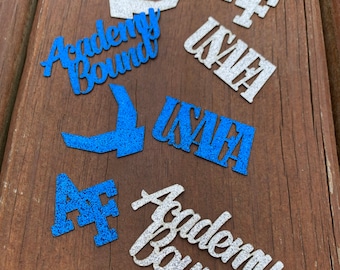 May include: A collection of six glitter-covered paper cutouts. The cutouts are in blue and silver. The cutouts include the words "Academy Bound", "AF", and "USAFA".